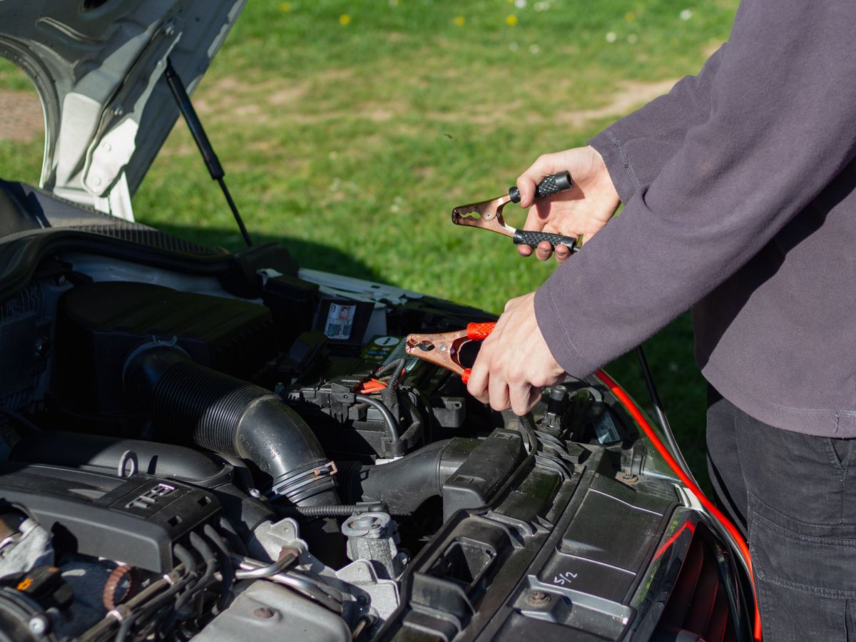 Hands clamping a set of jumper cables onto a car battery at an open hood.