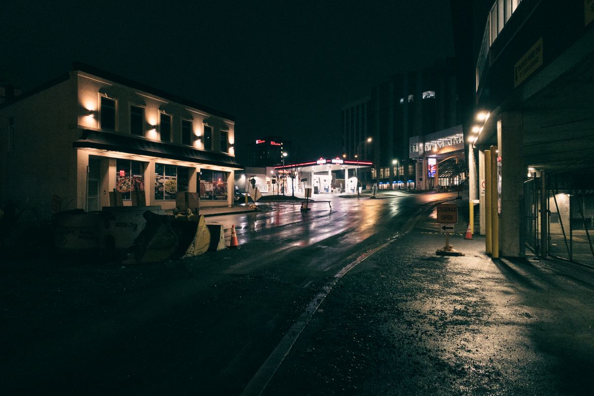 A wet empty street at night, a lit storefront on one side and a gas station glowing in the middle distance.