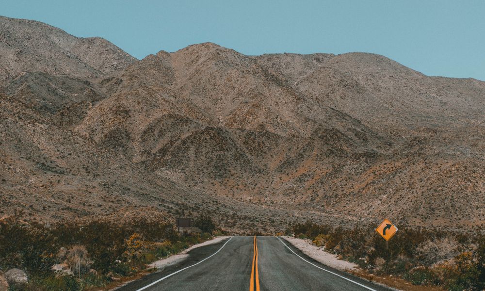 An empty two lane road with a yellow center line running toward dry high desert hills.