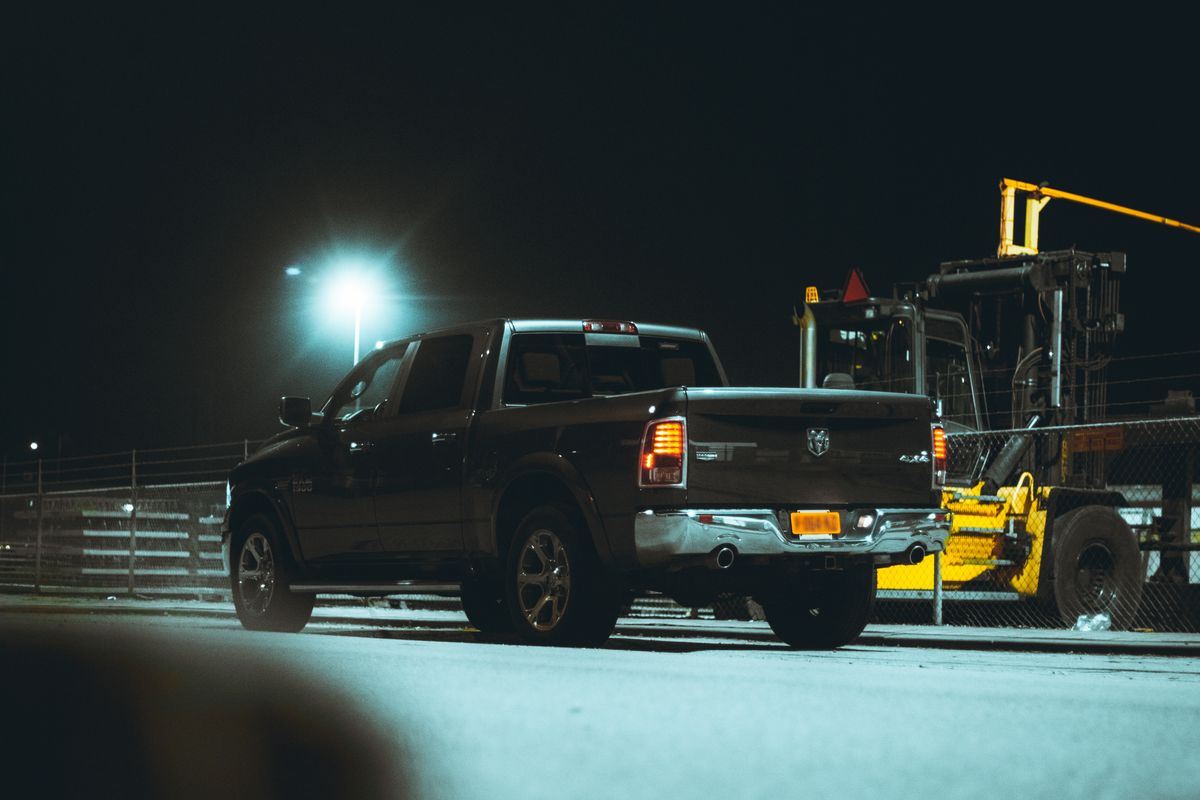 A dark pickup truck seen from behind at night in an industrial lot, taillights lit, a floodlight behind it.